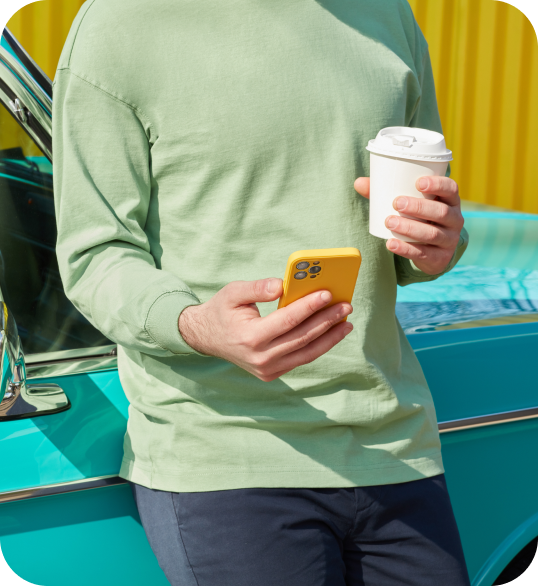 man in mint long sleeve shirt leaning against a turquoise car looking at his phone with a cup on coffee in left hand